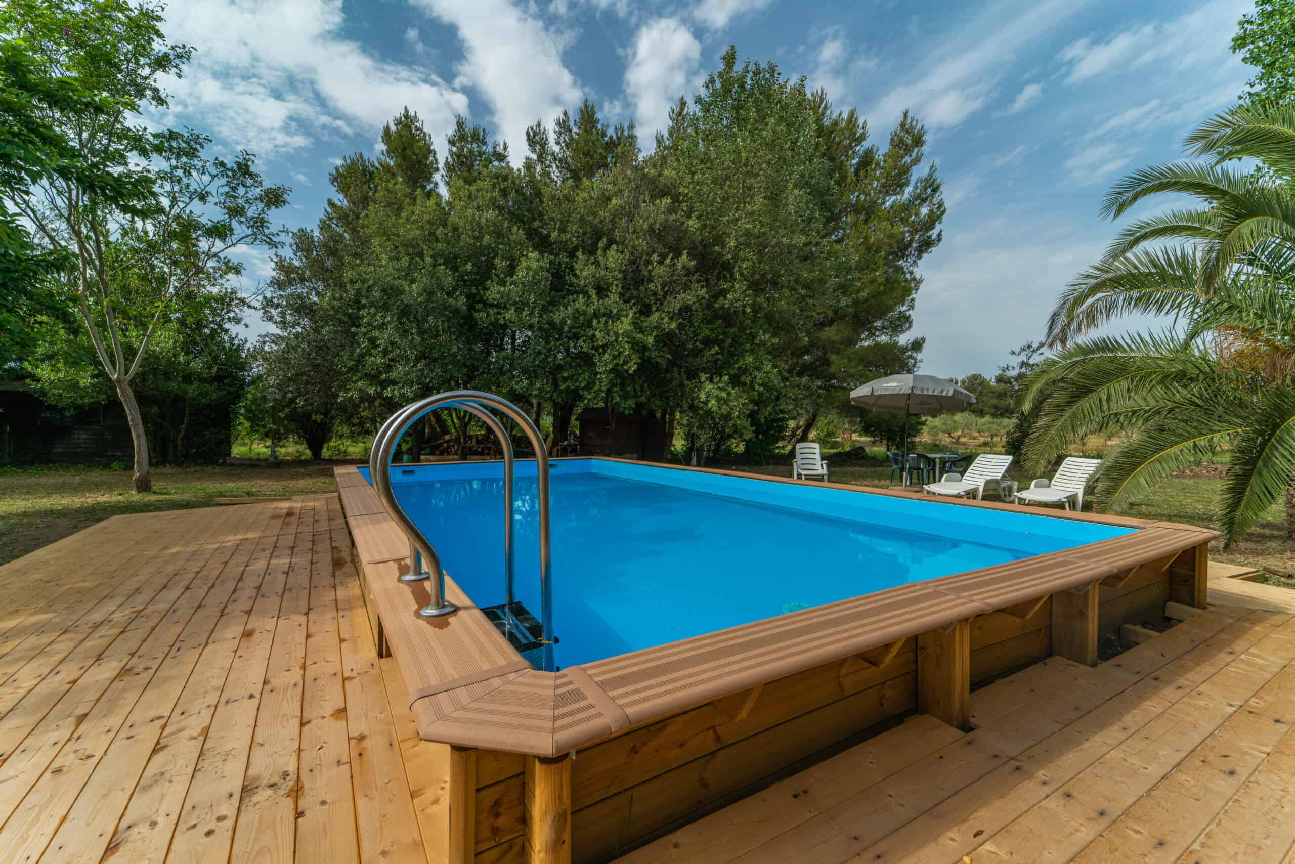 Piscina immersa nella natura con solarium in legno e vegetazione mediterranea.